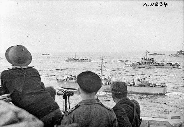 Black and white photograph. Three men face away from the camera, one in helmet, one in navy cap, one with no headwear. They look out over the ocean - many boats carrying men and equipment to the Dieppe Raid can be seen.